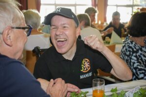 Mann mit Baseballcap lacht, reckt die Faust; schwarzes Poloshirt mit Schriftzug "MANUEL"; im Hintergrund Menschen an gedeckten Tischen.