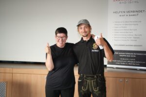 Zwei lächelnde Personen stehen nebeneinander und zeigen Daumen hoch, Mann trägt schwarzes Polo mit Schriftzug 'PATRICK', Rollup im Hintergrund