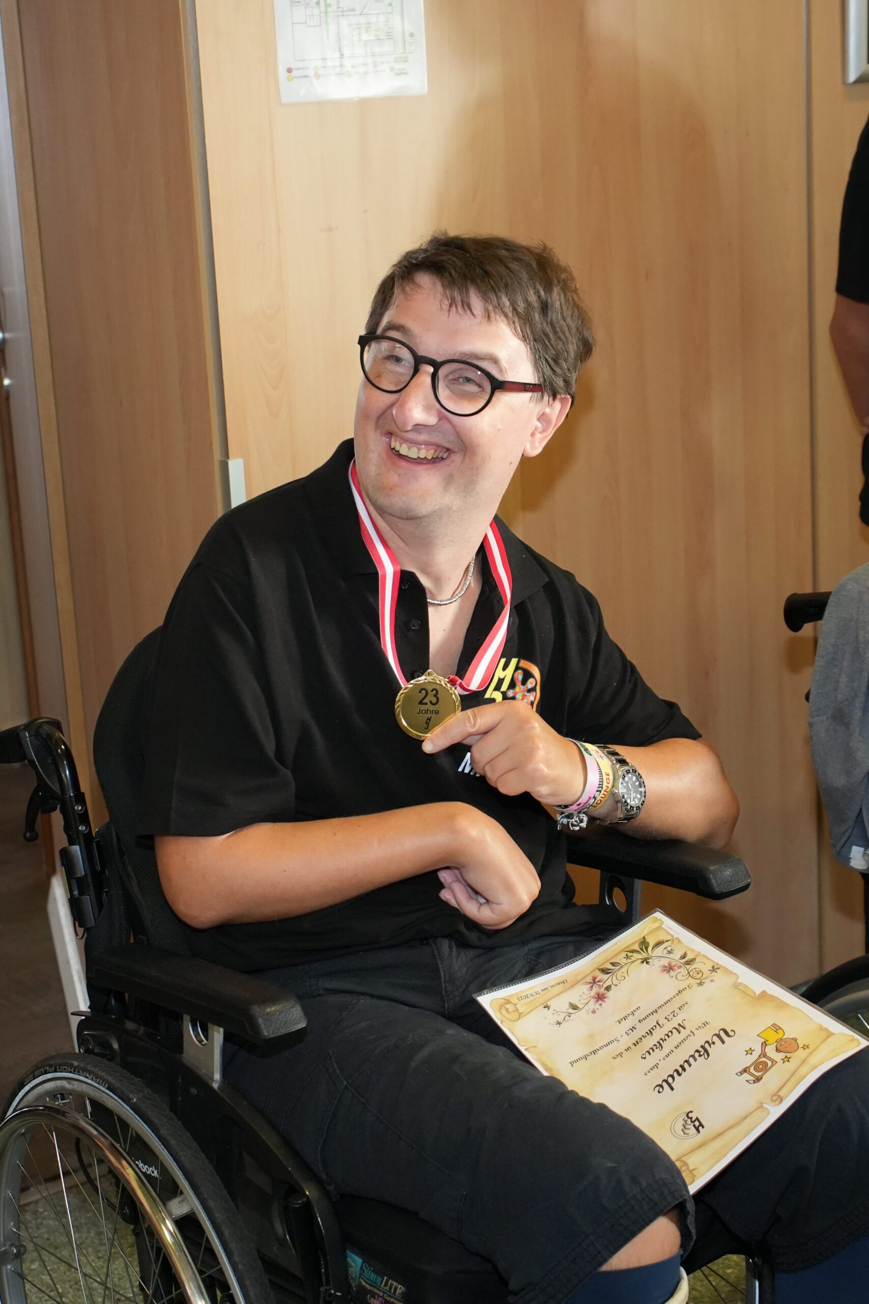 Lächelnder Mann im Rollstuhl mit Medaille (Aufschrift: "23 Jahre") und einer Urkunde auf dem Schoß.