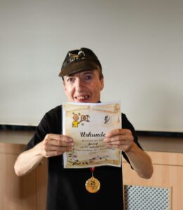 Mann hält eine Urkunde mit der sichtbaren Überschrift „Urkunde“ und eine Medaille mit der Aufschrift „25 Jahre“.